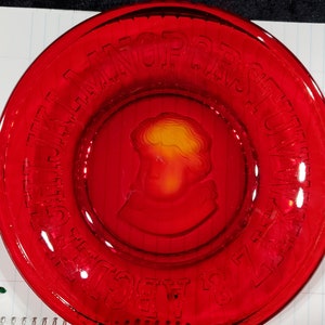 Puede incluir: Un plato de vidrio rojo con un diseño en relieve del alfabeto alrededor del borde. El centro del plato presenta una imagen en relieve de la cabeza de una persona.