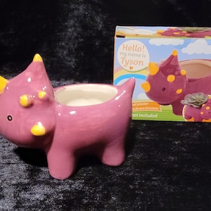 Puede incluir: Un macetero de dinosaurio de cerámica rosa con detalles amarillos. El macetero tiene forma de triceratops y tiene un pequeño agujero en la parte superior para plantar. La caja dice "Hello! My name is Tyson".