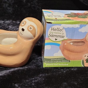 Puede incluir: Una maceta de cerámica con forma de perezoso en tonos beige y crema, con una cara sonriente. La maceta tiene una amplia abertura para plantas. La caja dice "Hello! my name is Sammie" y "animal planter".
