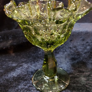 Könnte beinhalten: Ein hellgrünes Glas-Stielschale mit gerüschtem Rand und strukturiertem Muster. Die Schale steht auf einem kreisförmigen Fuß mit einem bauchigen Stiel. Das Glas hat einen leichten Gelbstich. Das Design erinnert an antike Glaswaren.