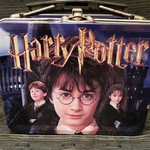 May include: A blue metal lunchbox with a Harry Potter design. The lunchbox features a picture of Harry Potter, Ron Weasley, and Hermione Granger from the first movie. The lunchbox has a black handle and a gold clasp.