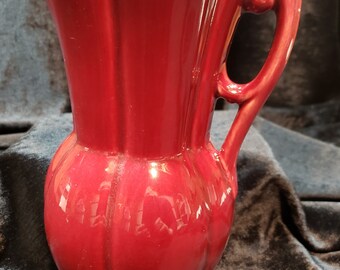 May include: A red ceramic pitcher with a fluted design and a handle. The pitcher has a rounded body and a flared lip.