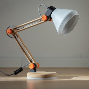 Puede incluir: Lámpara de escritorio moderna con estructura de madera clara, detalles en negro y naranja, y una pantalla gris. La lámpara tiene un brazo ajustable y una base redonda. El cable es gris y negro.