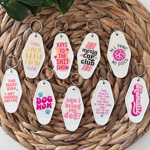 Peut inclure: Plusieurs porte-clés blancs de style motel avec des phrases et des graphiques variés. Les porte-clés incluent des phrases comme "Dog Mom", "Messy Car Club" et "All Panic No Disco". Chaque porte-clé a un anneau argenté.