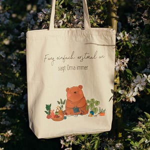 May include: A white canvas tote bag with brown straps. The bag has a brown bear illustration with the text "Fang einfach erstmal an sagt Oma immer".