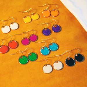 May include: A set of 10 pairs of colorful round earrings with gold hooks. The earrings are arranged in two rows on a yellow surface. The colors include white, yellow, orange, red, pink, purple, blue, green, and black.