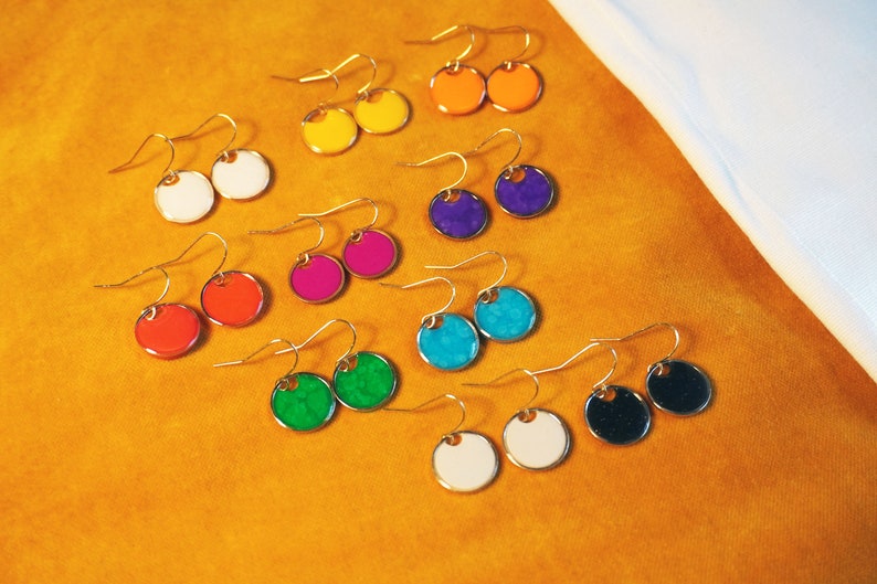 May include: A set of 10 pairs of colorful round earrings with gold hooks. The earrings are arranged in two rows on a yellow surface. The colors include white, yellow, orange, red, pink, purple, blue, green, and black.