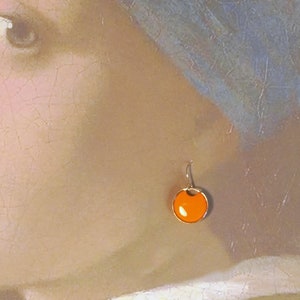 May include: A pair of orange and gold earrings with a round, slightly concave, orange stone. The earrings are hanging from a gold hook.
