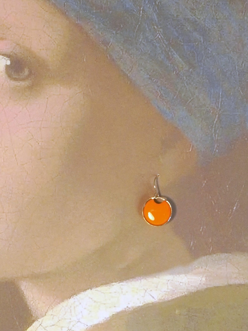 May include: A pair of orange and gold earrings with a round, slightly concave, orange stone. The earrings are hanging from a gold hook.