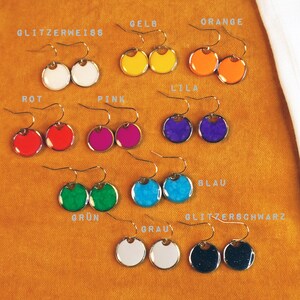 May include: A set of 10 pairs of earrings with different colored round discs. The colors include red, orange, yellow, green, blue, purple, pink, white, gray, and black. The earrings are labeled with the color names in German.