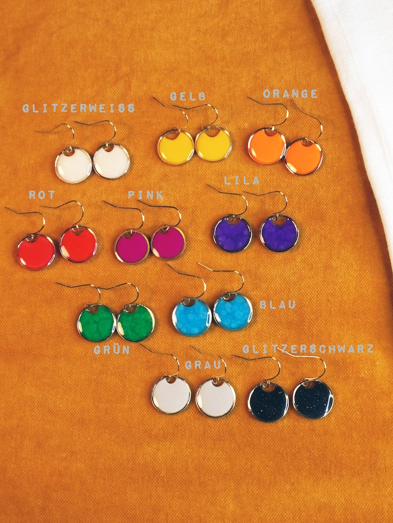May include: A set of 10 pairs of earrings with different colored round discs. The colors include red, orange, yellow, green, blue, purple, pink, white, gray, and black. The earrings are labeled with the color names in German.