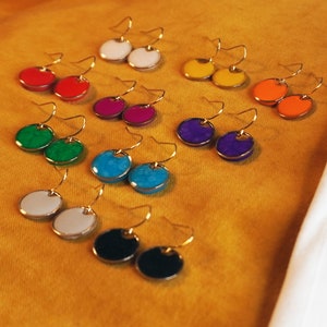 May include: Assortment of colorful round earrings with gold hooks. The earrings are arranged in pairs on a yellow surface. The colors include white, red, green, blue, purple, orange, and black.