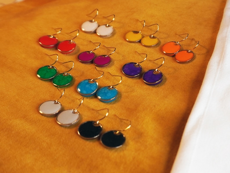 May include: Assortment of colorful round earrings with gold hooks. The earrings are arranged in pairs on a yellow surface. The colors include white, red, green, blue, purple, orange, and black.