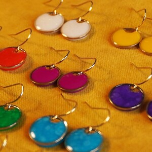 May include: Assortment of colorful earrings with gold hooks. The earrings feature round, painted metal discs in various colors, including red, white, yellow, purple, blue, and green.