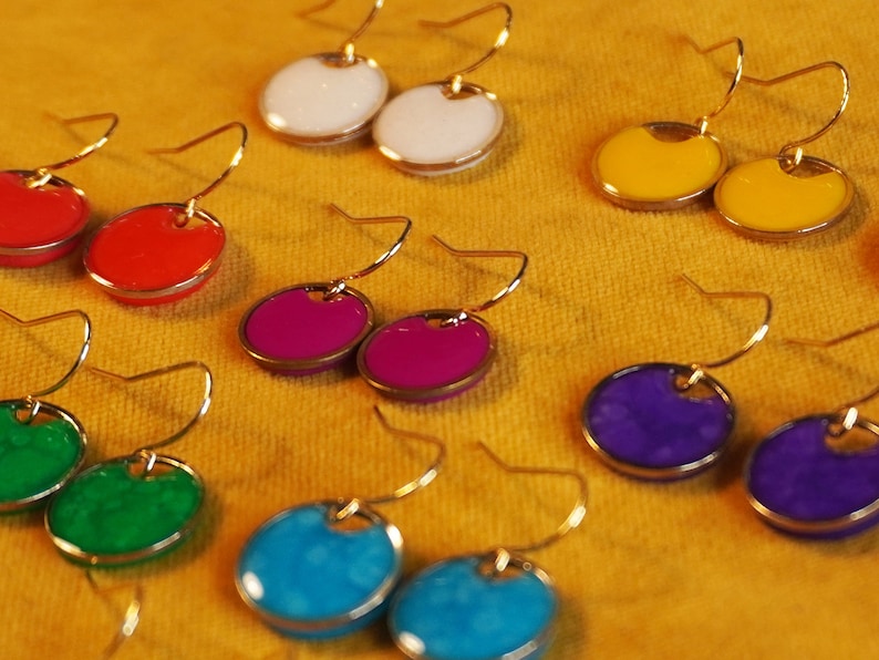 May include: Assortment of colorful earrings with gold hooks. The earrings feature round, painted metal discs in various colors, including red, white, yellow, purple, blue, and green.