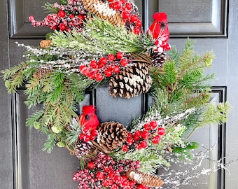 May include: A Christmas wreath with red berries, pine branches, pine cones, and two red cardinals. The wreath is decorated with faux snow.