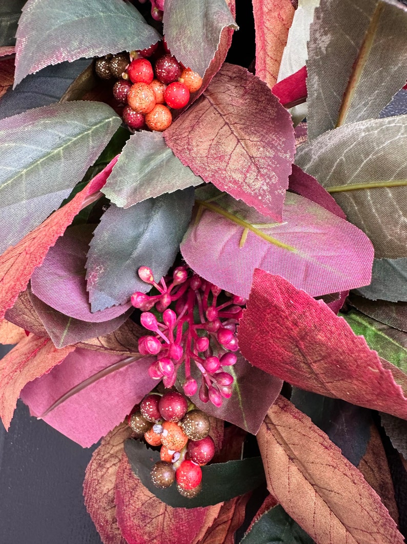 Pode incluir: Um close-up de uma coroa de outono com folhas bord&ocirc;, verdes e marrons e bagas vermelhas e marrons.