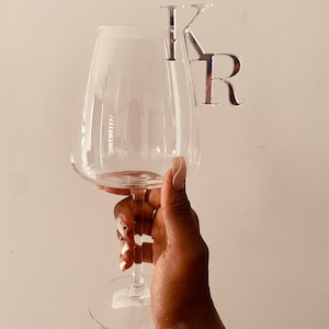 Könnte beinhalten: Ein klares Weinglas mit einem silbernen Metallmonogramm der Buchstaben "K" und "R", das am Rand befestigt ist. Das Glas wird vor einem schlichten weißen Hintergrund gehalten.