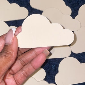May include: Several light beige cloud-shaped cutouts are displayed on a dark blue textured surface. One is held in a hand. The cutouts have a smooth finish, suitable for crafts or decoration. The cloud shapes are approximately 10 cm in length.