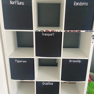May include: A white cube storage unit with nine compartments, each holding a black fabric bin. The bins are labeled "Nerf Guns", "Randoms", "Transport", "Figures", "DressUp", and "Creative". A toy gun rests on top.