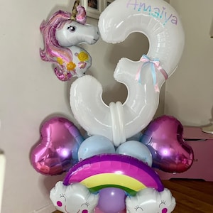 Puede incluir: Un arreglo festivo de globos para un cumpleaños. Incluye un globo blanco con el número 3 con el nombre "Amaiya", un unicornio rosa, globos en forma de corazón, un arcoíris y globos de nubes. Los globos son rosas, morados, azules y blancos.