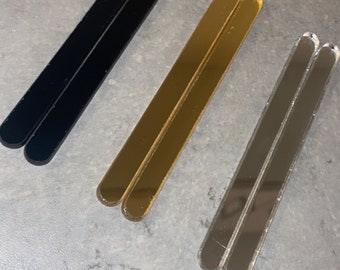 May include: Three pairs of acrylic stir sticks in black, gold, and silver. Each pair is rectangular with rounded ends, and they are arranged diagonally on a grey surface. These are ideal for mixing drinks.