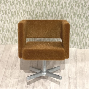 Puede incluir: Una silla giratoria marrón en miniatura tapizada con una base de metal cepillado. La silla tiene un respaldo bajo y reposabrazos, y el asiento es de forma cuadrada. La silla está diseñada para una casa de muñecas o un entorno en miniatura.