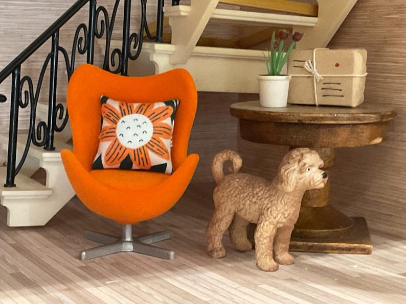 May include: An orange mid-century modern style chair with a floral pillow. A small brown dog figurine stands next to the chair. A small round table holds a plant and a wrapped package. The scene is set in a miniature interior.