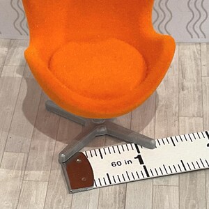 May include: An orange miniature chair with a silver base. The chair is approximately 1 inch wide. The chair is on a light gray and white patterned surface. The chair has a round seat and a curved back.