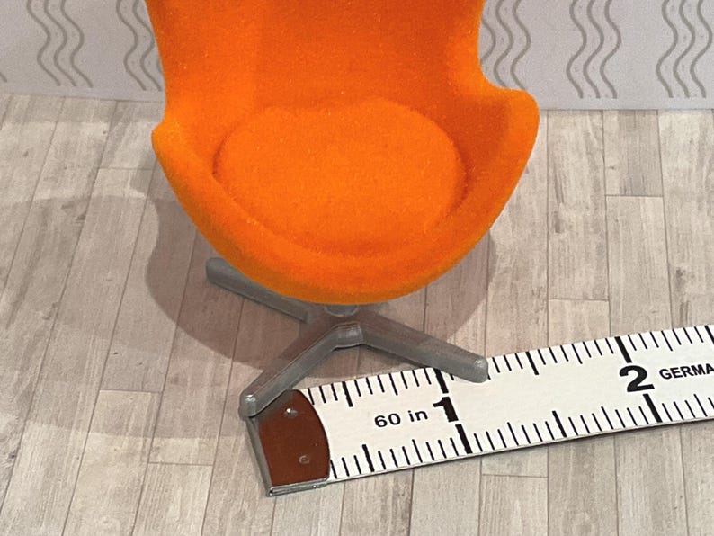 May include: An orange miniature chair with a silver base. The chair is approximately 1 inch wide. The chair is on a light gray and white patterned surface. The chair has a round seat and a curved back.