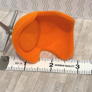 May include: An orange miniature chair with a silver metal base, next to a ruler. The chair is approximately 2 inches wide. The ruler is marked with the text "GERMAN QUALITY".