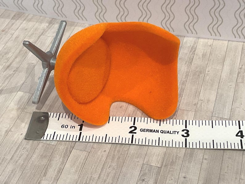 May include: An orange miniature chair with a silver metal base, next to a ruler. The chair is approximately 2 inches wide. The ruler is marked with the text "GERMAN QUALITY".