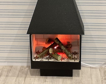 May include: A miniature black fireplace with a glowing red interior and faux logs. The fireplace has a star-patterned base and a black, angled top. A white cord extends from the base.