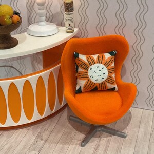 May include: An orange mid-century modern style chair with a floral pillow. The pillow has a white background with an orange flower design. The chair sits on a silver base. A white and orange table is in the background.