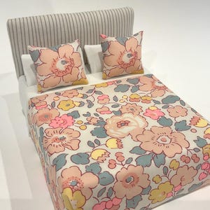 May include: Miniature bed with a striped headboard, white sheets, and two floral pillows. The bedspread features a repeating pattern of large pink, yellow, and blue flowers on a cream background. A charming, decorative piece.