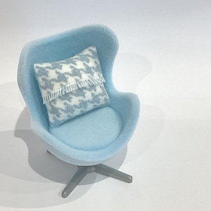 Op de afbeelding: Een miniatuur lichtblauwe stoel met een bijpassende zitting en rugleuning. Een kussen met patroon en franjes ligt op de stoel. De stoel heeft een draaivoet met drie poten. Het thema is miniatuurmeubels.