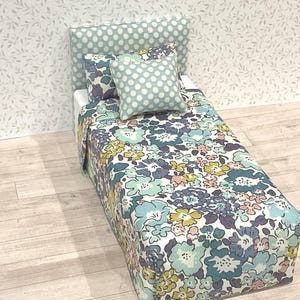 May include: A miniature bed with a floral patterned comforter and pillow. The bed frame and headboard are covered in a light blue fabric with white polka dots. The floral pattern includes blue, yellow, and purple flowers.
