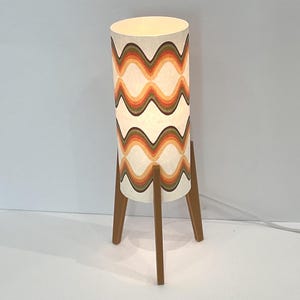 Op de afbeelding: Retro vloerlamp met een cilindrische kap met een golvend patroon in oranje, bruin en groen. De lamp staat op een houten driepoot, wat een mid-century modern gevoel geeft. De hoogte is ongeveer 100 cm.