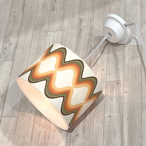 Könnte beinhalten: Zylindrischer Lampenschirm mit Retro-Design. Der Schirm hat einen cremefarbenen Hintergrund mit einem sich wiederholenden Wellenmuster in Orange, Grün und Braun. Eine weiße Deckenleuchte und ein Kabel sind befestigt.