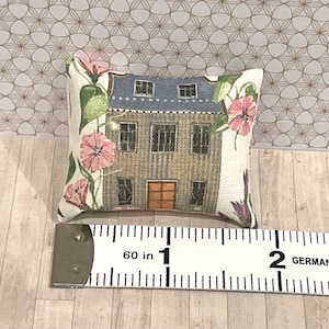 Könnte beinhalten: Ein Miniatur-Dekokissen mit einer detaillierten Illustration eines Gebäudes mit blauem Dach und oranger Tür. Das Kissen ist mit rosa floralen Akzenten verziert und misst etwa 2,5 cm in der Länge.