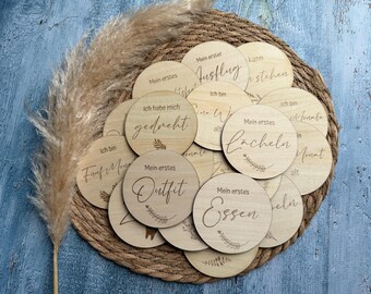 May include: Wooden milestone discs with German text, including "Mein erstes Lächeln" (My first smile), "Mein erstes Outfit" (My first outfit), and "Ich habe mich gedreht" (I turned over).