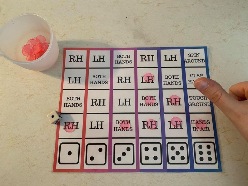 Pode incluir: Um tabuleiro de jogo com instru&ccedil;&otilde;es e dados. O tabuleiro tem uma grade com instru&ccedil;&otilde;es como "RH", "LH" e "Both Hands". Uma x&iacute;cara cont&eacute;m pe&ccedil;as de jogo rosa. Um dado mostra um &uacute;nico ponto.