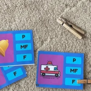 May include: A set of blue and purple flashcards with images of a bell, a campfire, and an ambulance. Each card has the letters P, MF, and F printed on it. There are also wooden clothespins on a beige carpet.