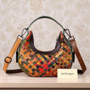 May include: A crescent-shaped handbag featuring a multi-colored woven leather exterior in shades of red, orange, yellow, green, and blue. It has a dark brown leather trim, a green top handle, and a tan adjustable shoulder strap. A card reads SacDesigns.