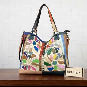 Peut inclure: Un sac cabas en cuir multicolore présentant un motif floral en patchwork avec diverses nuances de bleu, vert, rose, jaune et marron. Le sac comprend une bande verticale centrale avec des clous métalliques, deux anses et un petit panneau indiquant SacDesigns.
