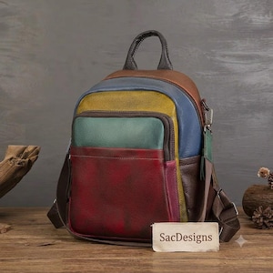 Op de afbeelding: Een veelkleurige leren rugzak met een handvat aan de bovenkant en verstelbare schouderbanden. De tas heeft horizontale panelen in bruin, blauw, geel, groen en donkerrood. Een rechthoekig label met de tekst SacDesigns is aan de voorzijde bevestigd.