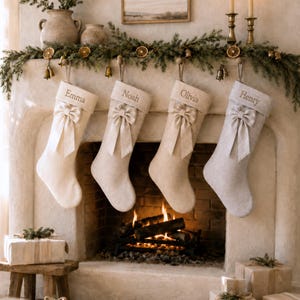Può includere: Quattro calze di Natale personalizzate appese in fila, ognuna realizzata in tessuto di colore chiaro con un fiocco decorativo sulla parte superiore. Le calze sono ricamate con i nomi Emma, Noah, Olivia e Henry in un carattere semplice.