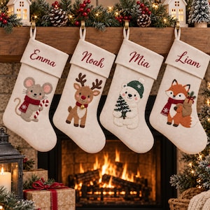 Op de afbeelding: Vier crèmekleurige kerstsokken van vilt hangen op een rij. Elke sok is gepersonaliseerd met een in rood schrift geborduurde naam: Emma, Noah, Mia en Liam. De sokken zijn versierd met vilten applicaties van een muis, een rendier, een ijsbeer en een vos, die winteraccessoires zoals sjaals en mutsen dragen.