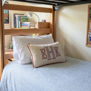 May include: A rectangular cream-colored throw pillow featuring a monogram with the letters EHK embroidered in light blue with a dark red outline. The pillow has a dark red piped edge and rests on a bed with a blue and white striped bedspread.