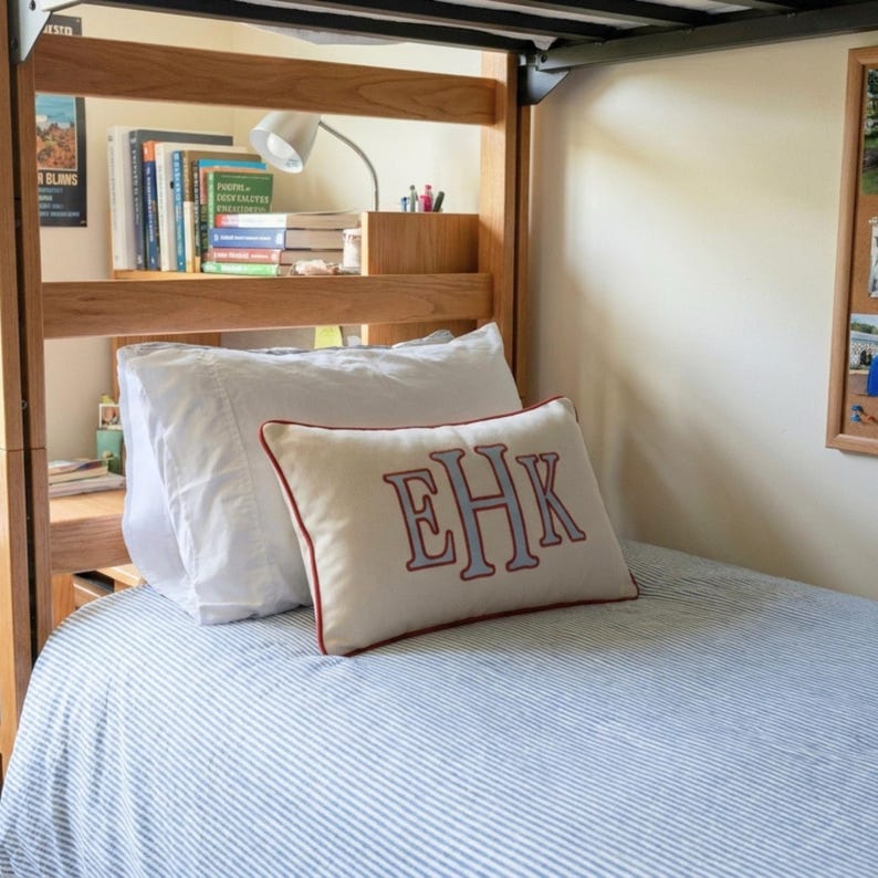May include: A rectangular cream-colored throw pillow featuring a monogram with the letters EHK embroidered in light blue with a dark red outline. The pillow has a dark red piped edge and rests on a bed with a blue and white striped bedspread.